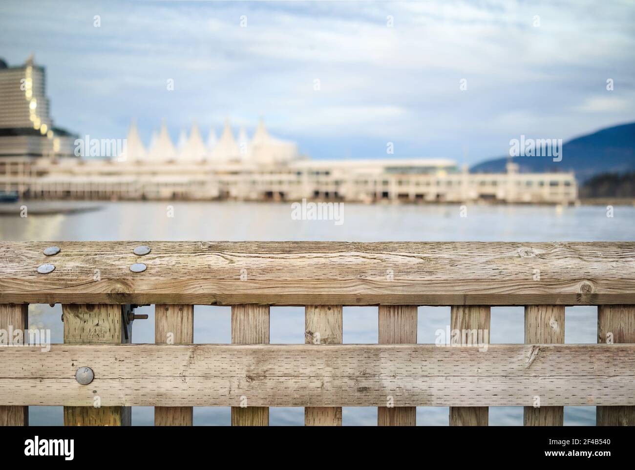 Ringhiera in legno con edifici poco nitidi sul lungomare e sfondo dell'oceano e cielo nuvoloso. Edificio sfocato e astratto del Porto di Vancouver Foto Stock