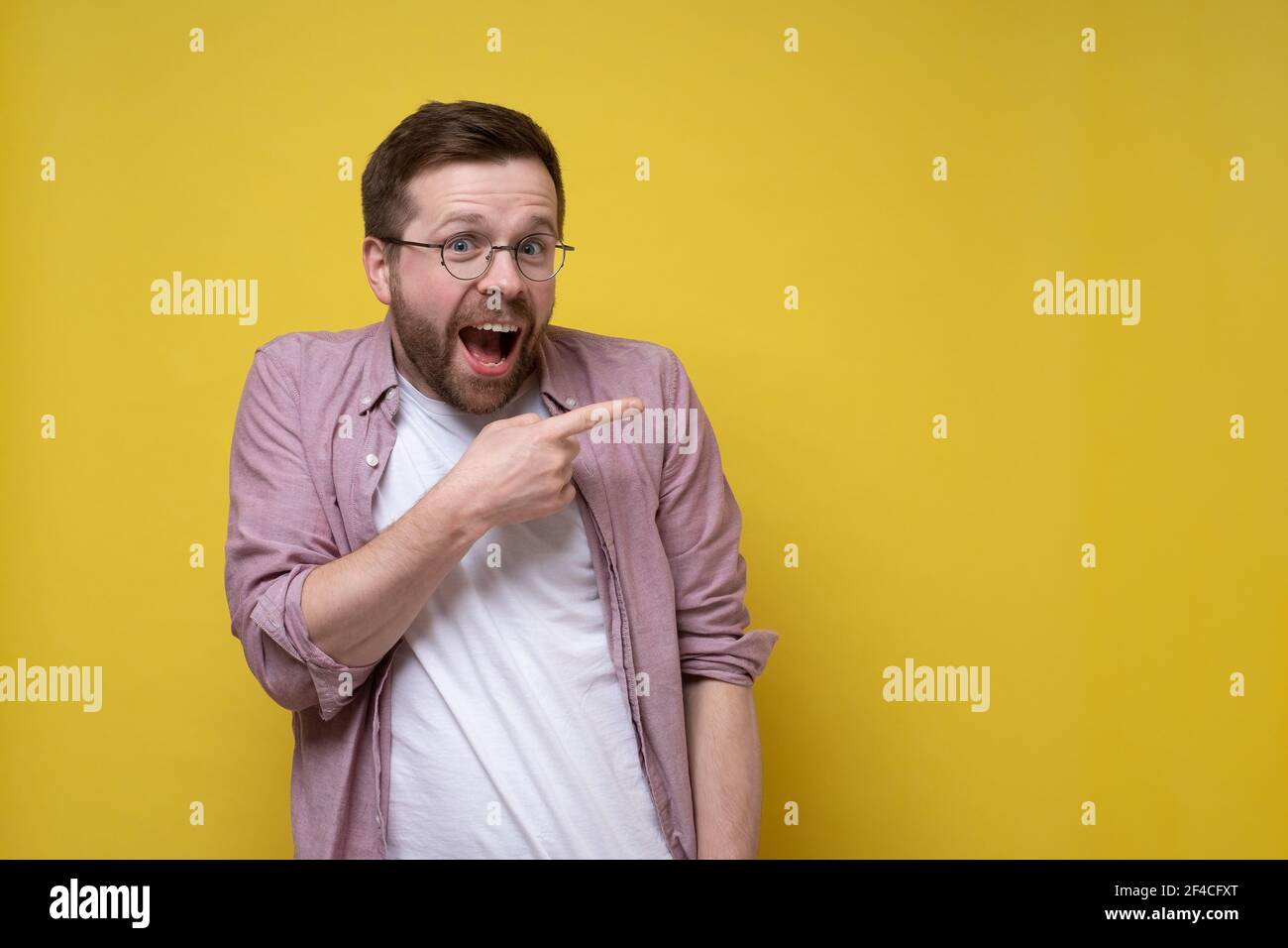 Uomo sorpreso e affascinante in una camicia rosa e gli occhiali puntano il dito a lato e guarda felicemente la fotocamera. Spazio di copia. Foto Stock