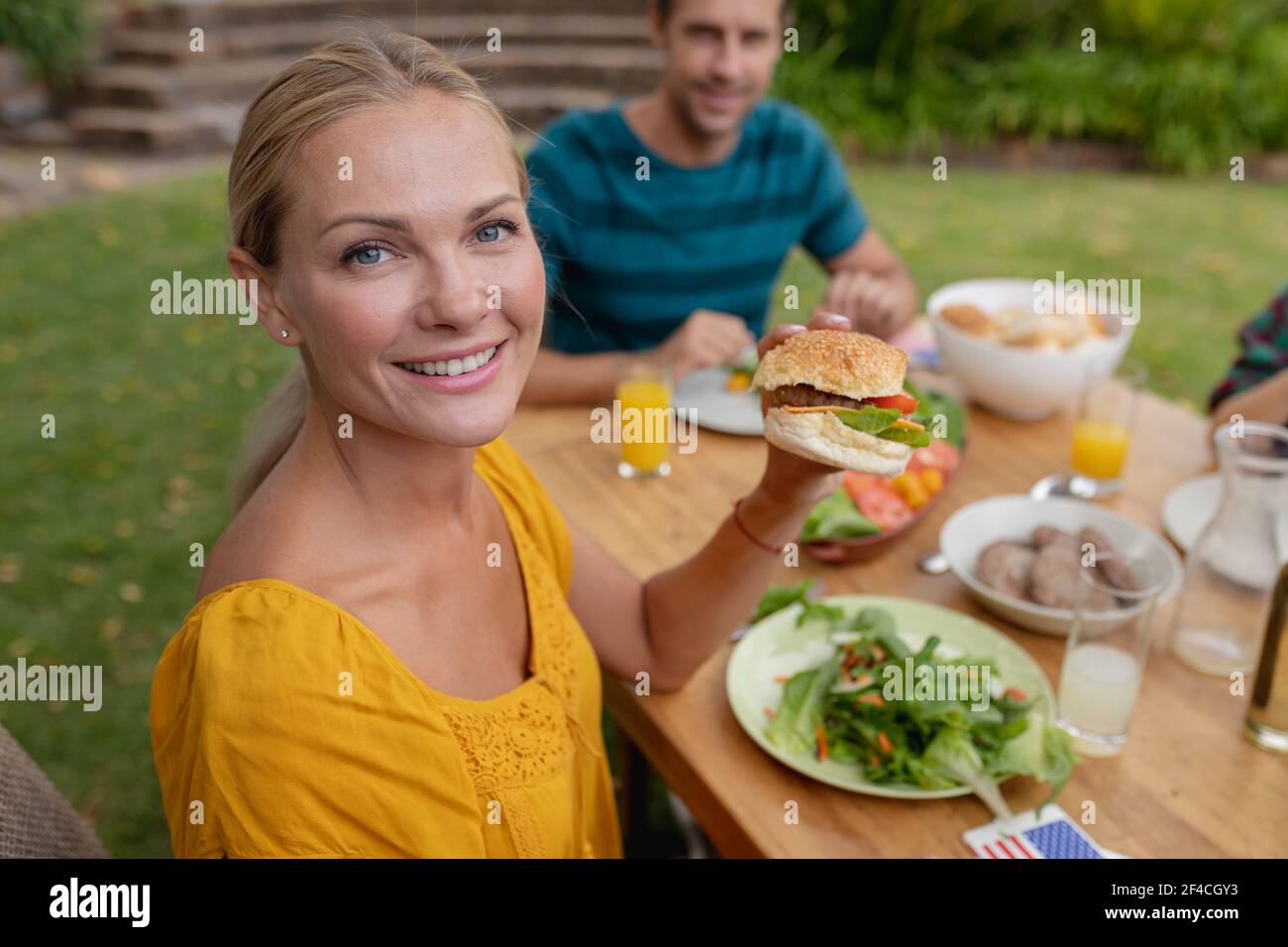 Ritratto di sorridente caucasica donna che tiene hamburger mangiare con famiglia in giardino Foto Stock