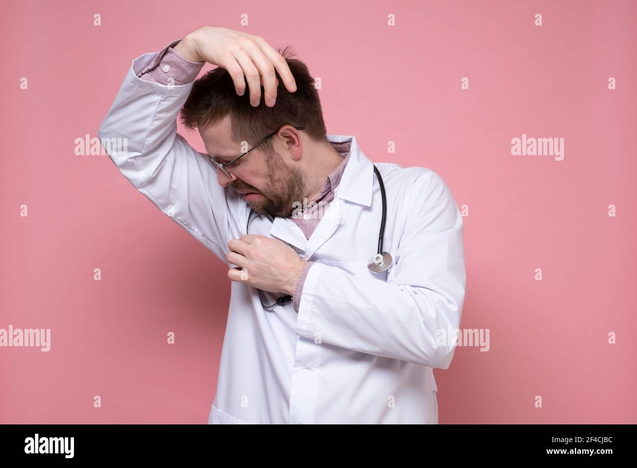 Il medico maschile in un cappotto bianco, con uno stetoscopio, sniffa le sue ascelle, irritato dal problema del sudore e dello stenching del corpo. Foto Stock