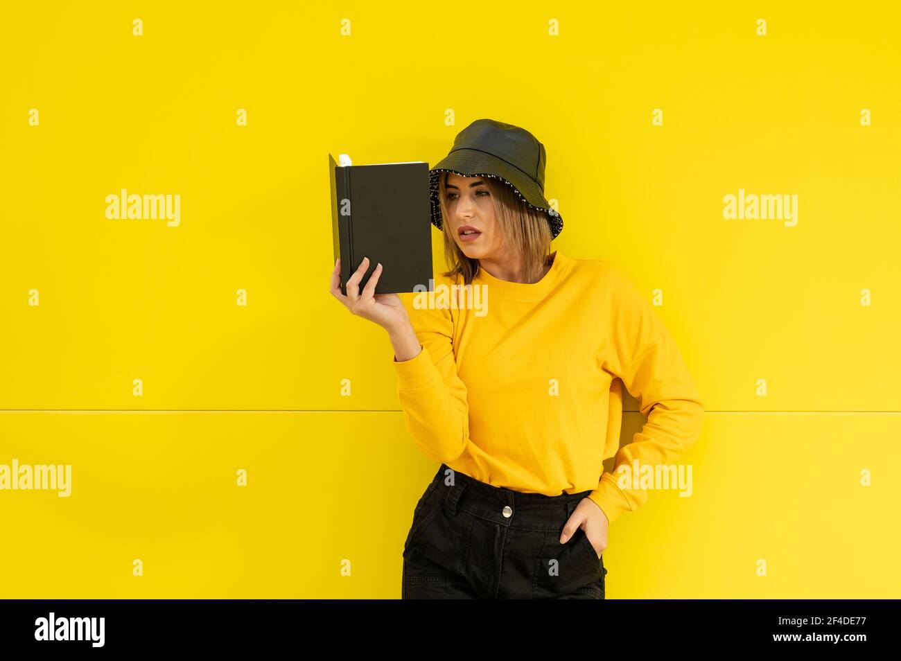 Un primo piano di una giovane donna caucasica in giallo con un cappello nero che legge un libro Foto Stock