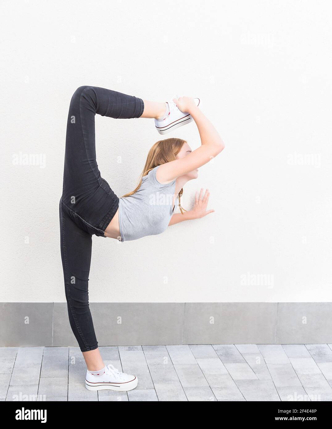 la ragazza dai capelli rossi nel profilo in posa del balletto sollevando la gamba sopra la testa con sneakers bianchi, pantaloni neri e t-shirt grigia su sfondo grigio chiaro Foto Stock