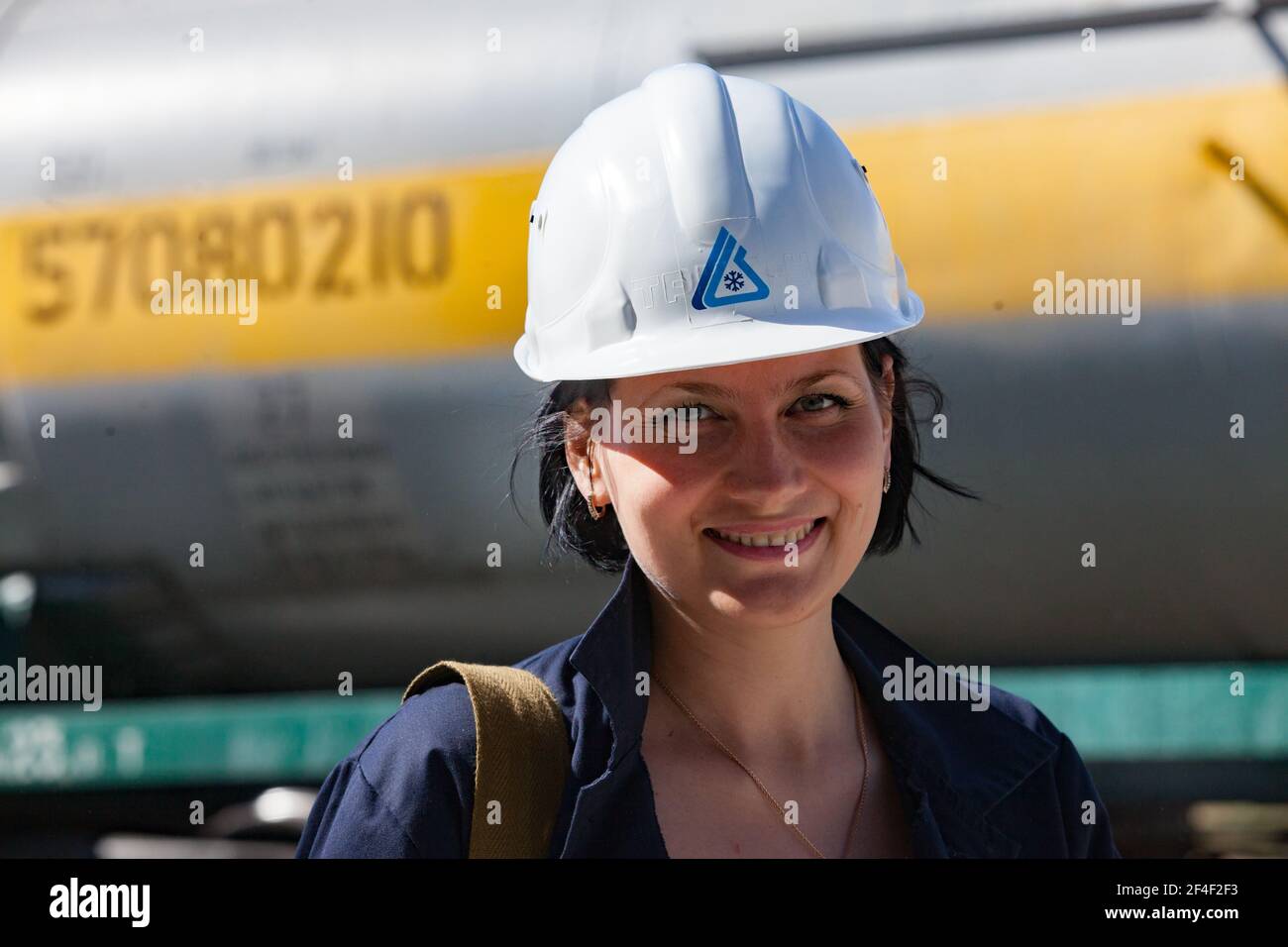 Impianto chimico caustico. Attraente giovane ingegnere ragazza in hardHat bianco che posa e sorridente sullo sfondo del carro armato ferroviario. Foto Stock