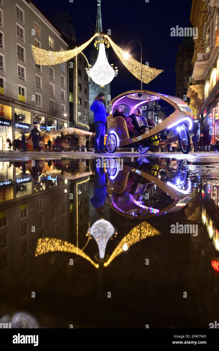 Un veicolo turistico in carrozza per visitare la storica città di Vienna con un grande lampadario come decorazioni natalizie riflesse in un pozze d'acqua. Foto Stock