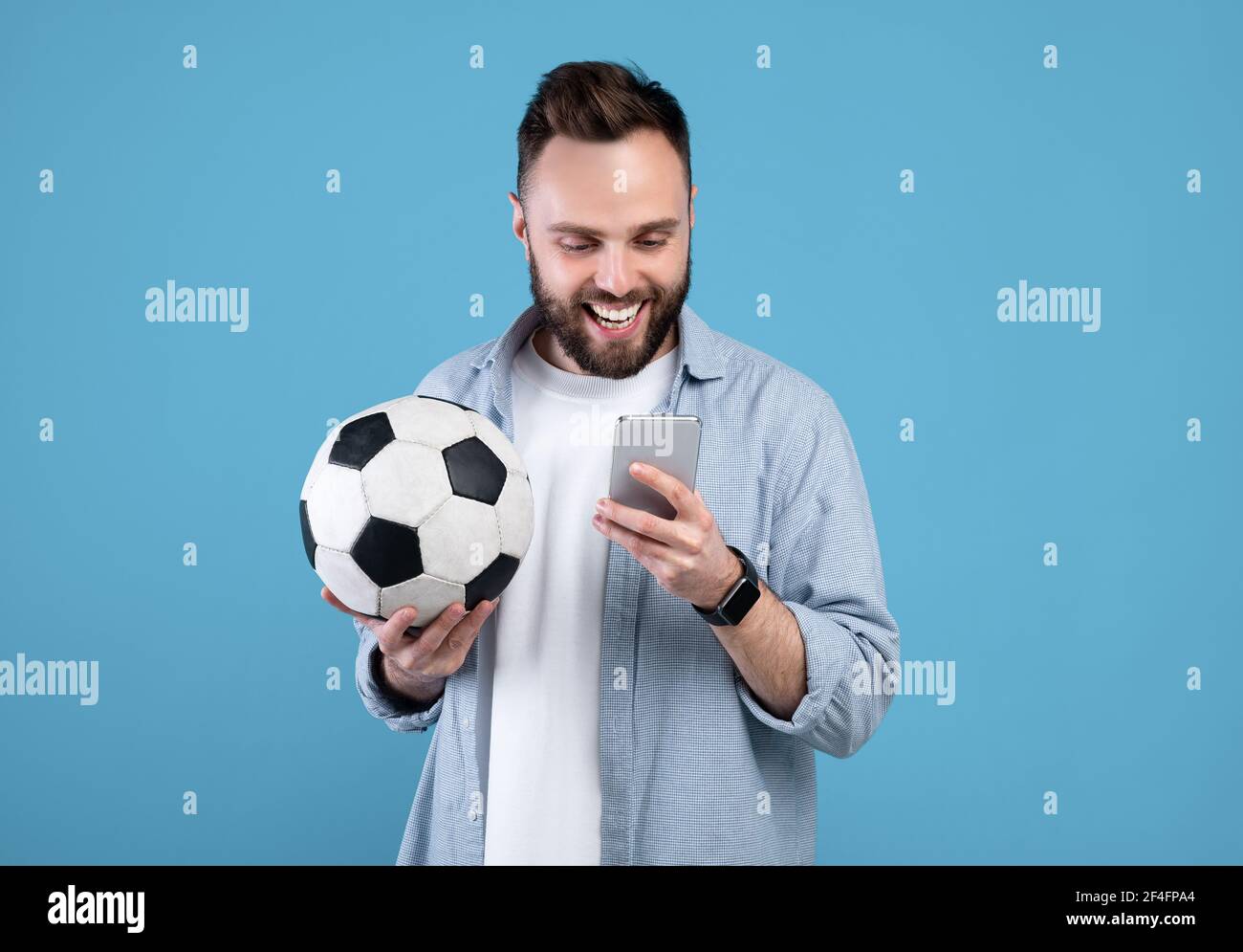 Felice giovane ragazzo con palla di calcio utilizzando smartphone, vincendo scommesse sportive, roting per la sua squadra preferita su sfondo blu Foto Stock