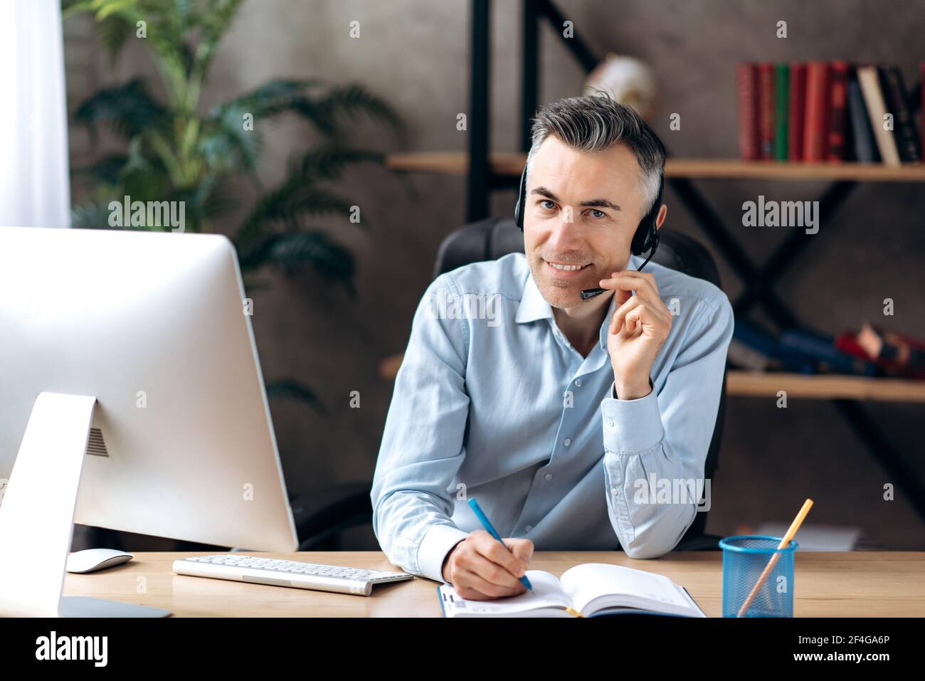 Un uomo d'affari adulto intelligente di successo si siede al moderno ufficio formale indossare e cuffia, comunica con un cliente o colleghi prende appunti in un notebook, guarda la fotocamera e sorridendo Foto Stock