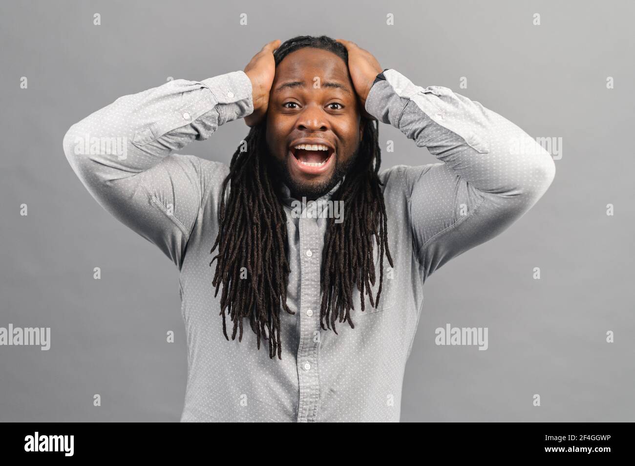 Ritratto di un giovane afro-americano scioccato con dreadlock che indossa una camicia guardando la macchina fotografica a sorpresa, stordito con una storia incredibile, tenendo le mani sulla testa, emozioni di espressioni umane Foto Stock