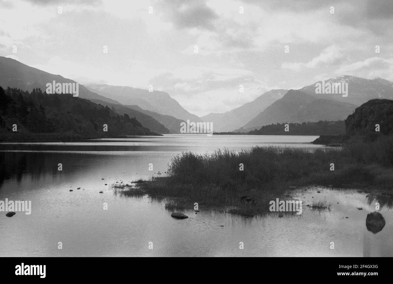 Anni 60, mattina presto e attraverso le acque di un lago, una vista a distanza del paesaggio di montagna di Snowdonia, Galles. Foto Stock