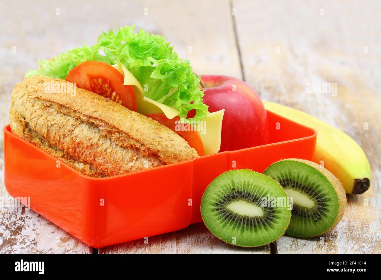 Una sana scatola da pranzo scolastica contenente un rotolo di formaggio a grani interi, mela, banana e kiwi Foto Stock