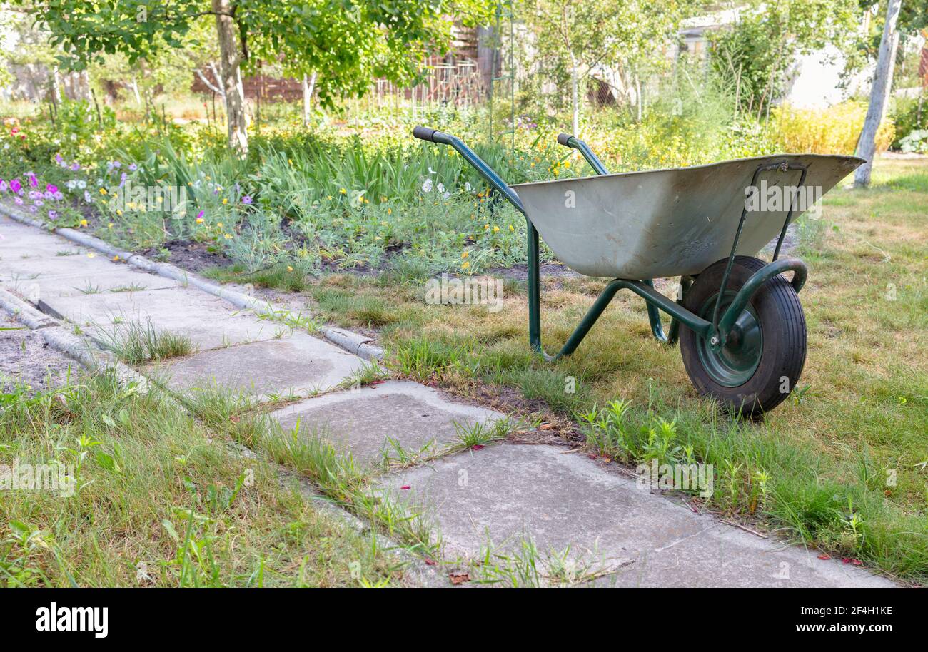 Un carro da giardino sorge su un prato verde vicino ad un vecchio sentiero in cemento sullo sfondo di un giardino estivo in sfocatura. Foto Stock