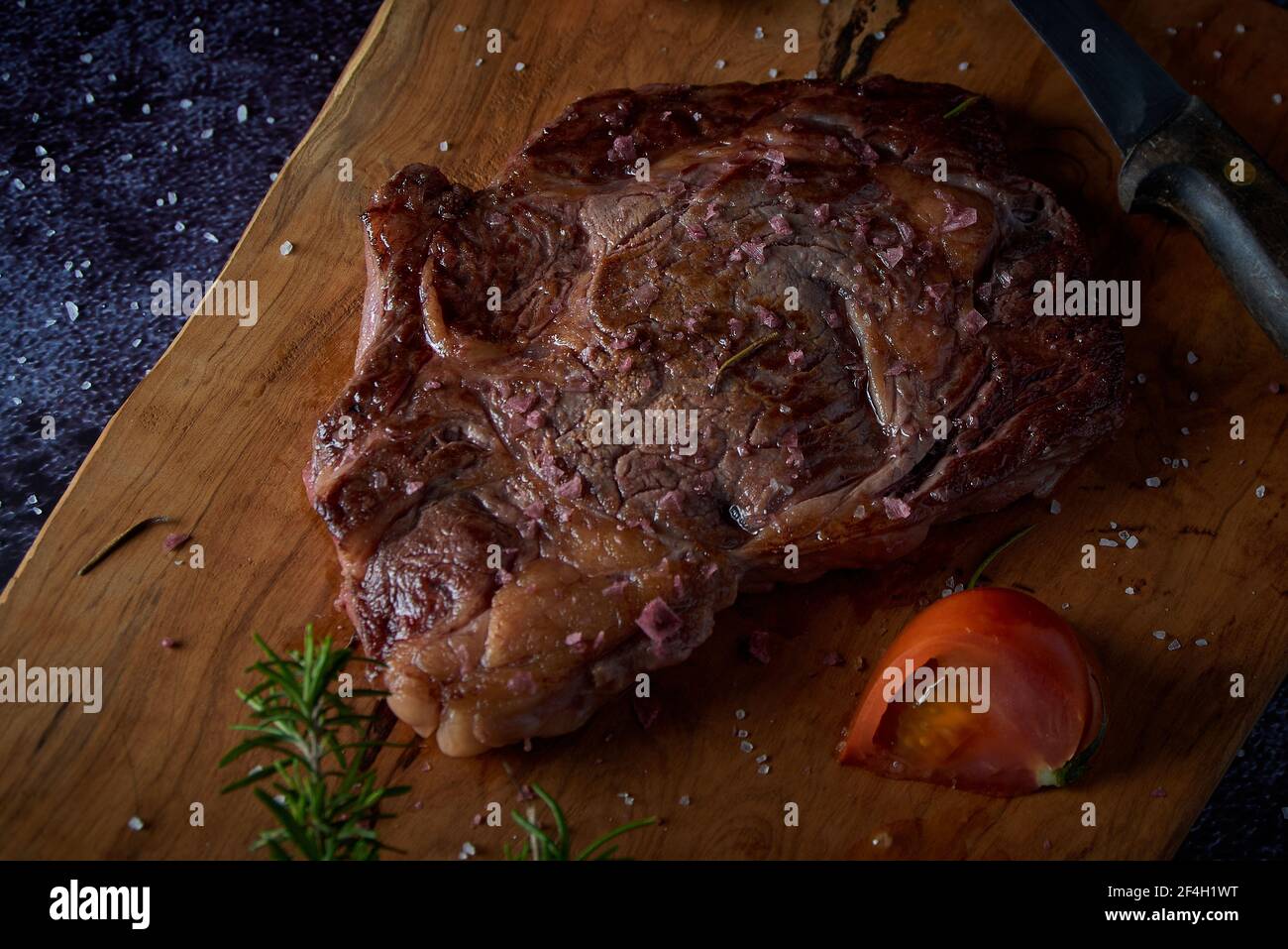 Bistecca di manzo alla griglia su tavola di legno con sale e altri condimenti su una superficie scura. Bistecca pronta per mangiare. Cibo scuro. Carne, pomodoro, pepe, limone AN Foto Stock