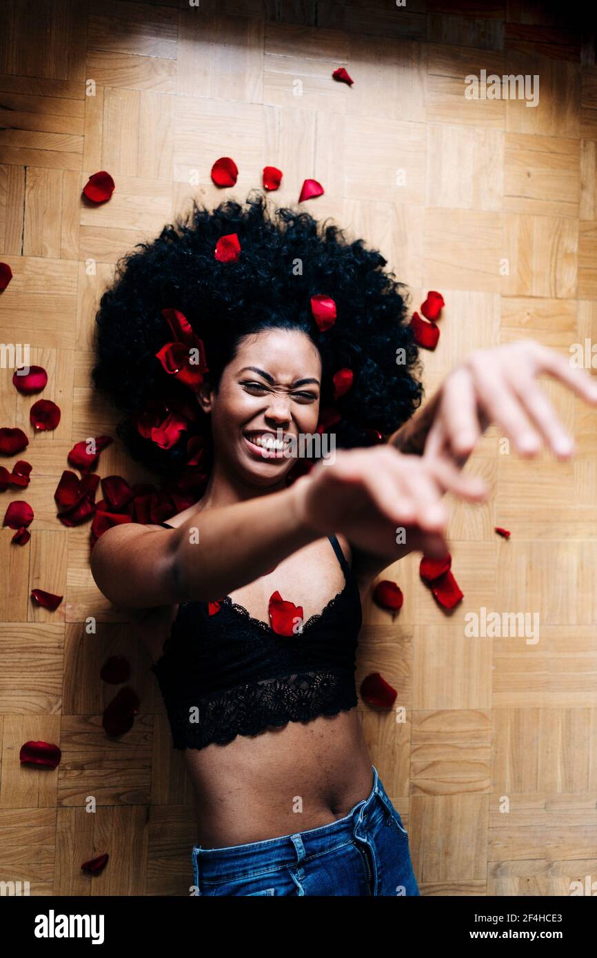 Vista dall'alto della romantica e allegra donna afroamericana sdraiata pavimento con petali di rosa rossa con occhi chiusi Foto Stock