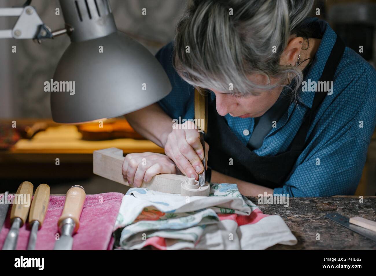 Donna concentrata con ornamento di taglio dei capelli grigi su pegbox vicino alla lampada mentre si crafting violino in officina Foto Stock