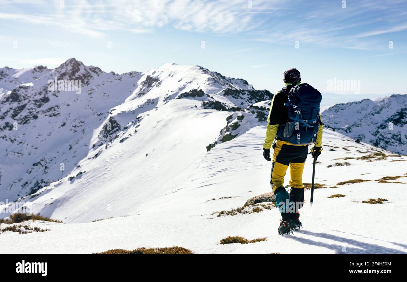 Vista posteriore di un arrampicatore irriconoscibile che cammina su un pendio di neve coperta catena montuosa rocciosa in tempo soleggiato Foto Stock