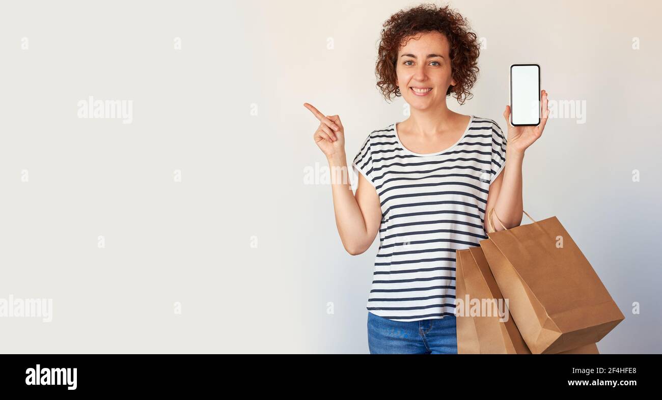 La donna caucasica dai capelli ricci punta al lato mentre tiene il suo telefono cellulare e alcune borse della spesa contro un isolato sfondo bianco Foto Stock