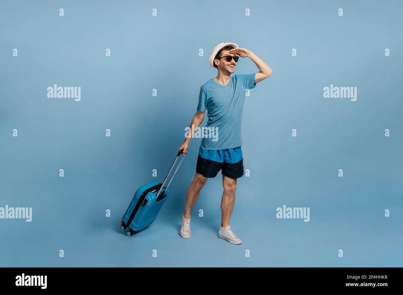 Un gioioso ragazzo caucasico a lunghezza intera in un cappello e occhiali da sole, vestito in abito estivo, tiene una valigia blu, guarda in lontananza, felice di una vacanza attesa a lungo, sfondo blu isolato Foto Stock