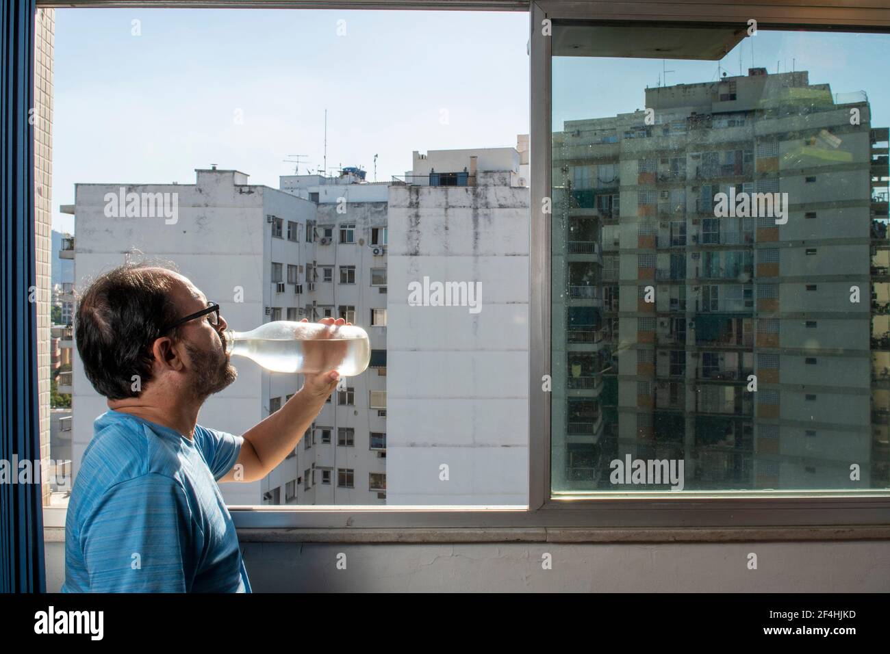 uomo che beve acqua dalla bottiglia alla finestra durante pandemia Foto Stock