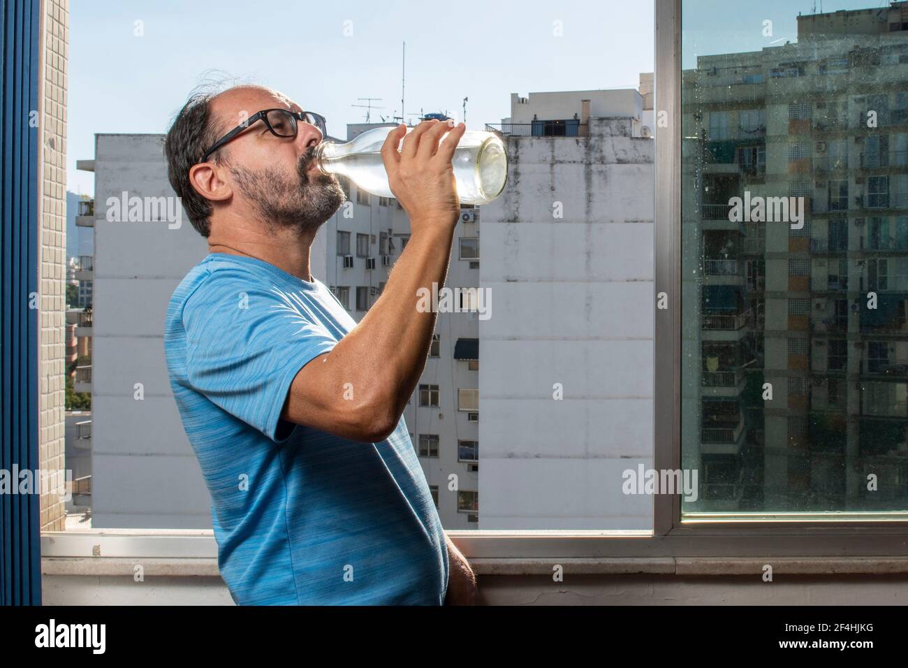 uomo che beve acqua dalla bottiglia alla finestra durante pandemia Foto Stock