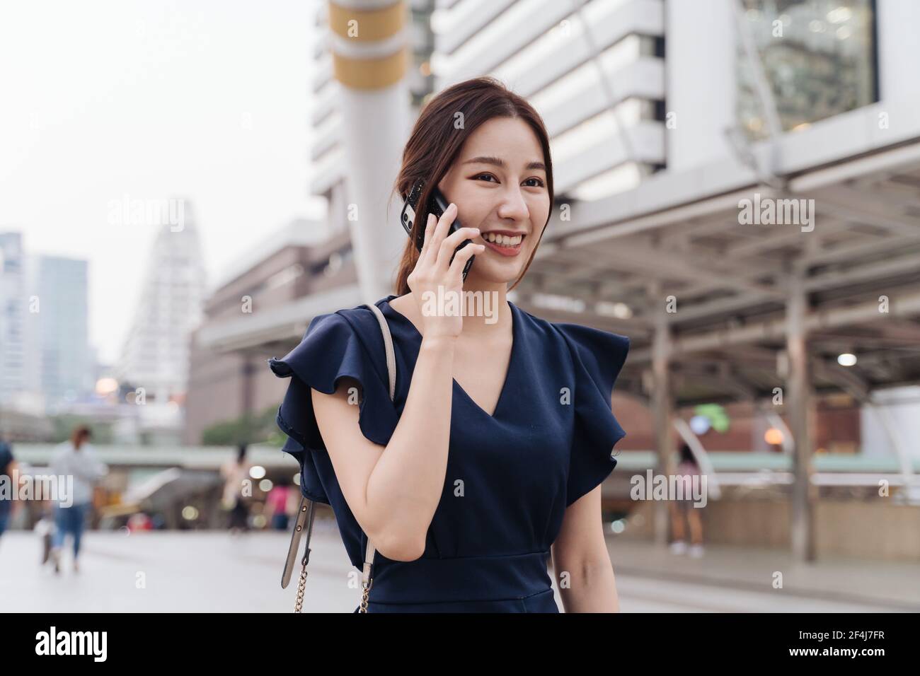 Ritratto di bella e sorridente giovane donna asiatica in casual abbigliamento che parla con un amico con uno smartphone mentre cammini in città via Foto Stock