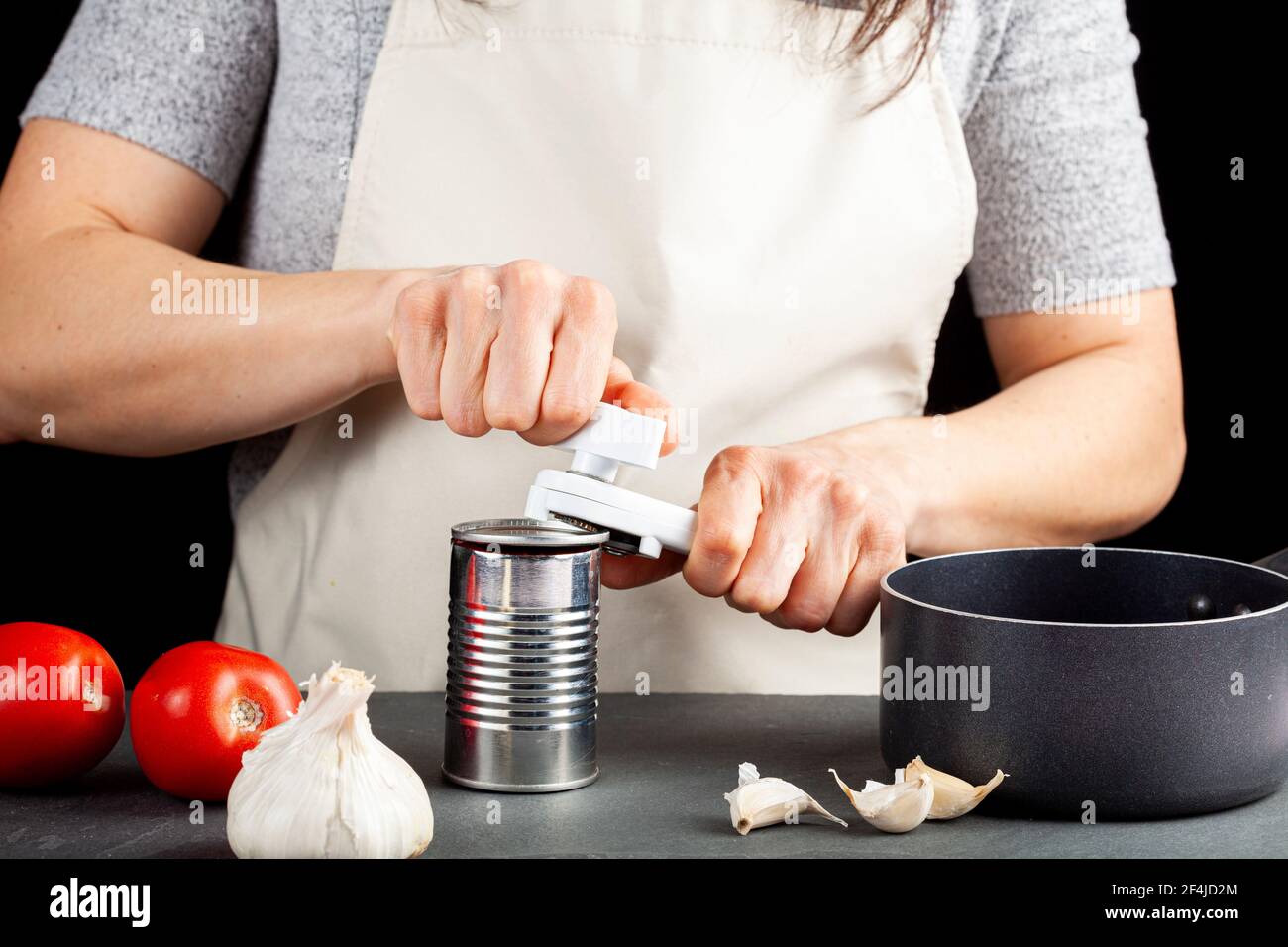 Una donna sta aprendo con attenzione una lattina di pasta di pomodoro su un banco da cucina utilizzando un apriboccatole in plastica bianca. Sta preparando un pasto per cui usa b Foto Stock