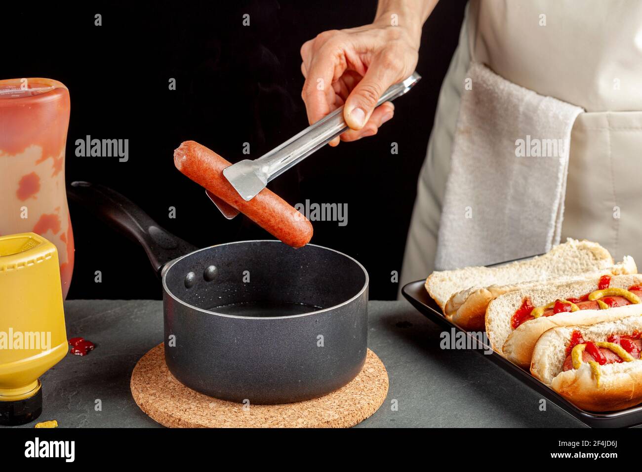 Una donna caucasica sta prendendo una salsiccia calda di cane bollita fresco da una pentola usando il tong di metallo per metterlo in un panino caldo del cane. Hot pot è su un sughero Foto Stock
