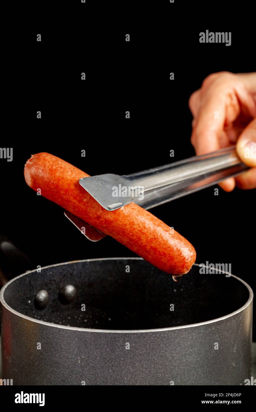 Una donna caucasica sta sollevando un singolo cane caldo dall'acqua bollente usando una tong di metallo. Si vede una pentola da cucina antiaderente e uno sfondo nero. Homema Foto Stock