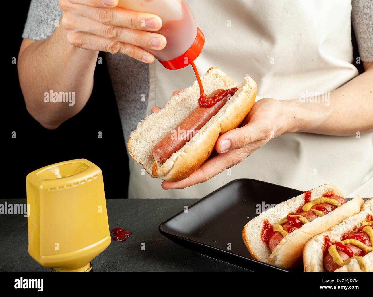 Una donna caucasica sta preparando panini caldi del cane. Sta spremendo il ketchup di pomodoro dalla bottiglia di plastica sopra ad una salsiccia di manzo all'interno di un mazzolino caldo del cane. Altri Foto Stock