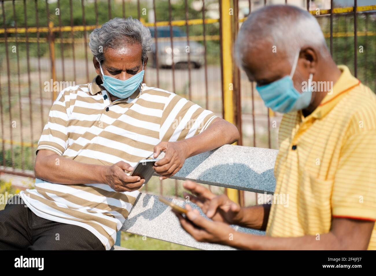 Vista laterale di due uomini anziani con maschera medica uso telefono cellulare mentre si è seduti al parcheggio mantenendo la distanza sociale concetto di coronavirus covid Foto Stock