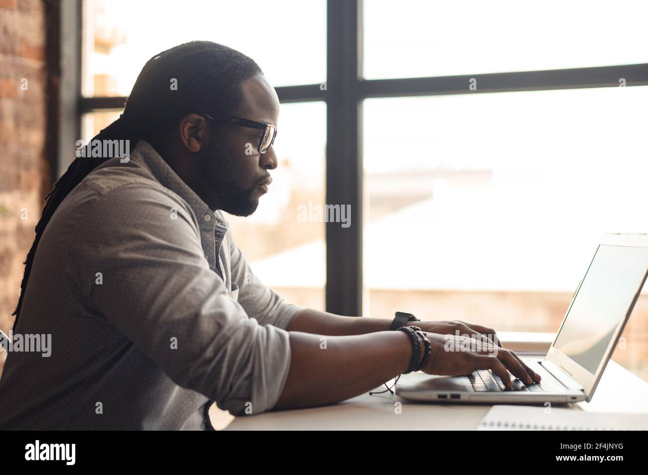 Vista frontale di occupato giovane afroamericano maschio autore studente o imprenditore in occhiali con dreadlock seduto alla scrivania digitando sul laptop, lavorando sul progetto, la scrittura di un articolo o blog Foto Stock