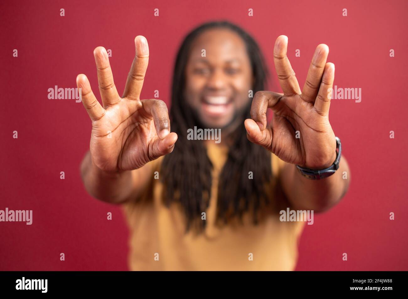 Uomo afroamericano con dreadlock isolato sulla parete rossa guardando la macchina fotografica, che mostra il segno giusto con entrambe le mani, gesturing e sorridente, felicità, concetto di pace e benessere, fuoco selettivo sulle mani Foto Stock