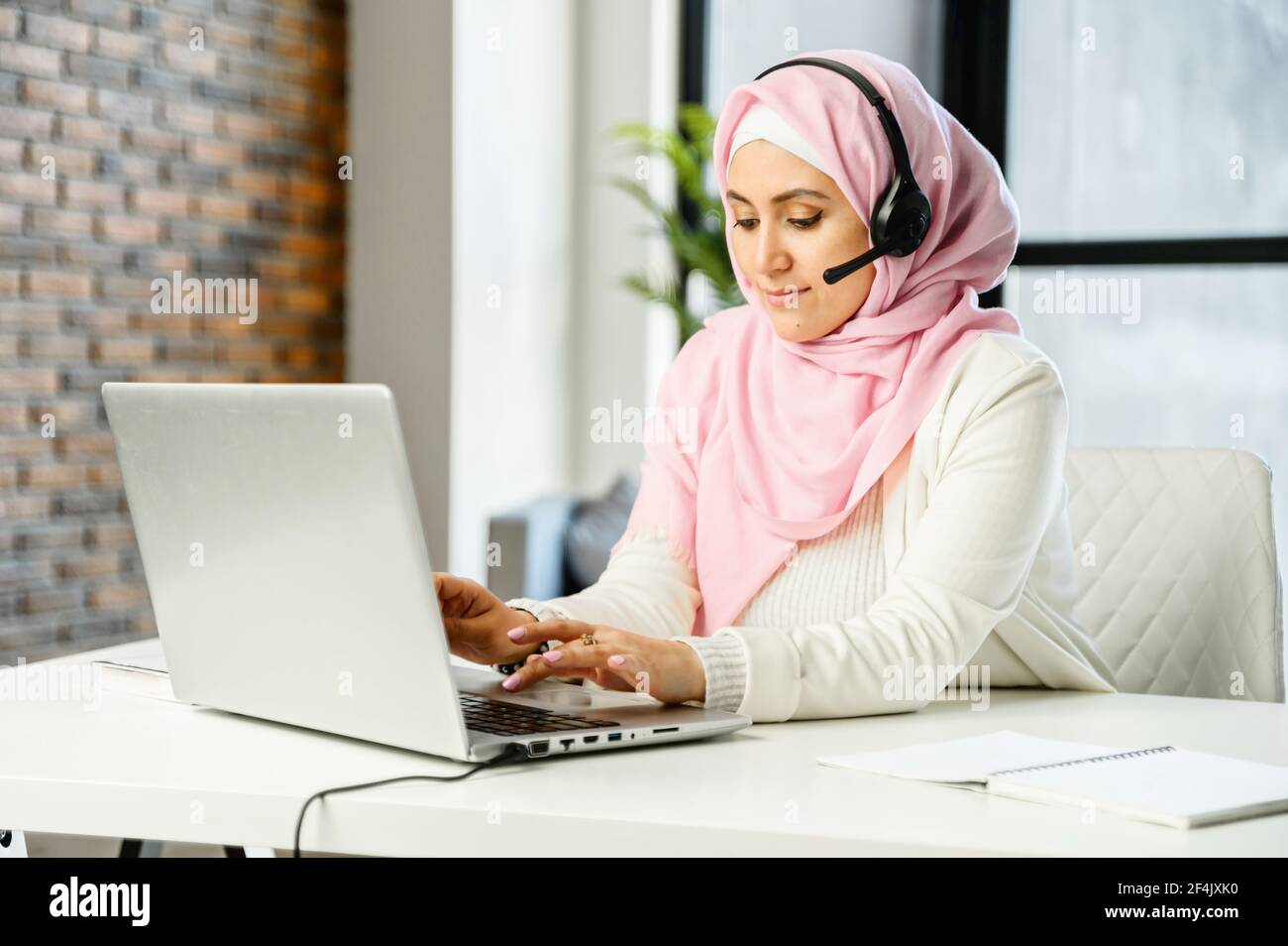 Giovane musulmano arabo misto-razza rappresentante del servizio clienti femminile in hijab e cuffie seduta alla scrivania in ufficio, digitando e-mail sul portatile, cercando o navigando in internet, rispondendo al cliente Foto Stock