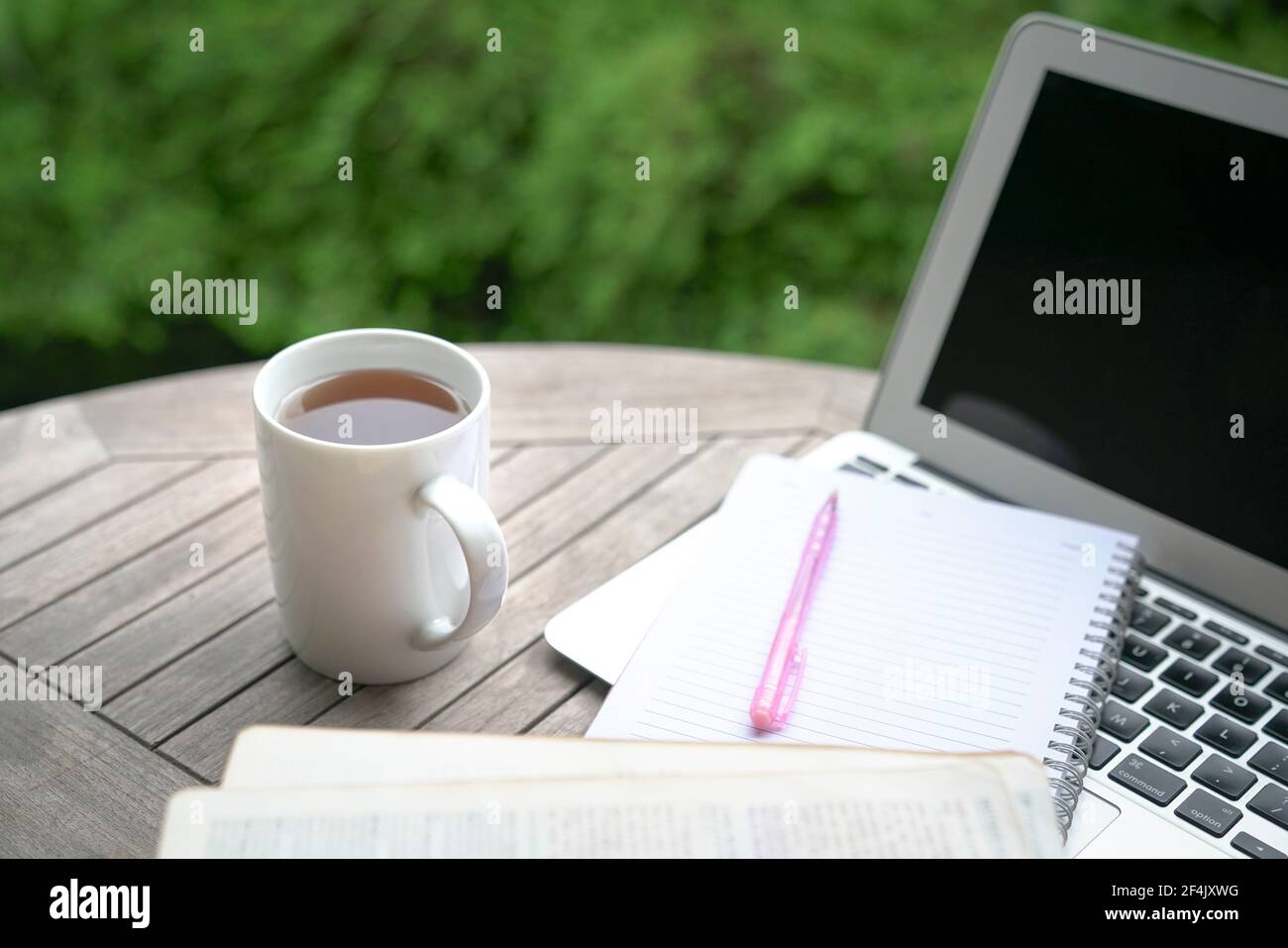 Tazza di caffè, blocco note, libro aperto, penna e computer portatile sul tavolo con piante verdi sullo sfondo. Foto Stock
