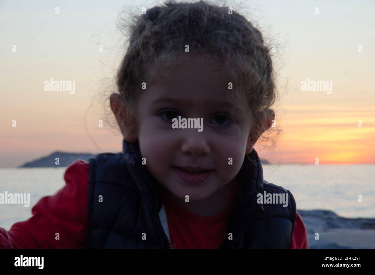 chubby piccola ragazza con capelli biondi fluttering guarda la macchina fotografica parte anteriore della vista del tramonto, volto e guancia a fuoco selettivo Foto Stock