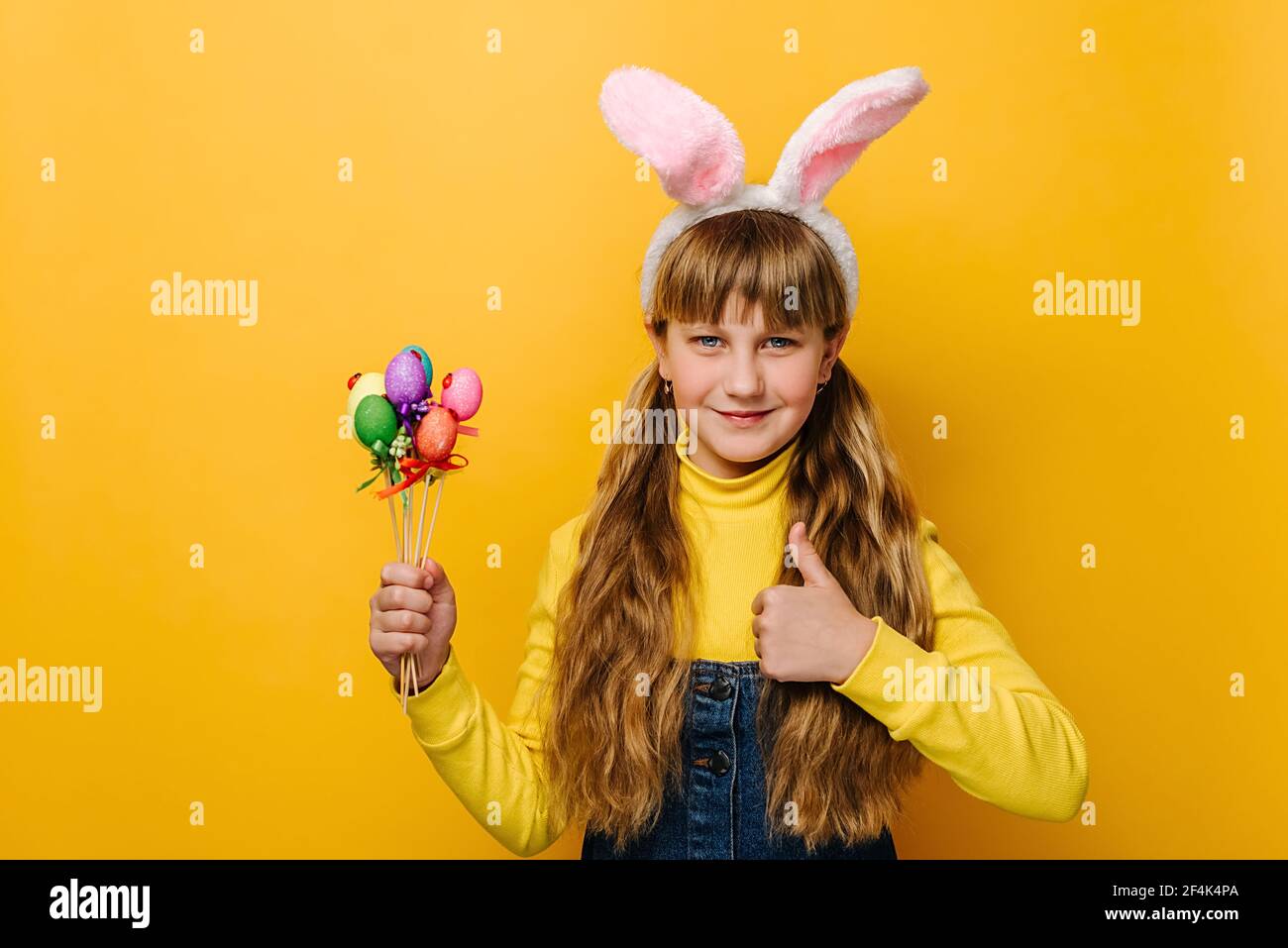 La bambina allegra indossa orecchie di lepre rosa, tenendo piccole uova colorate, mostrando il pollice in su, come gesto, dimostrando approvazione Foto Stock