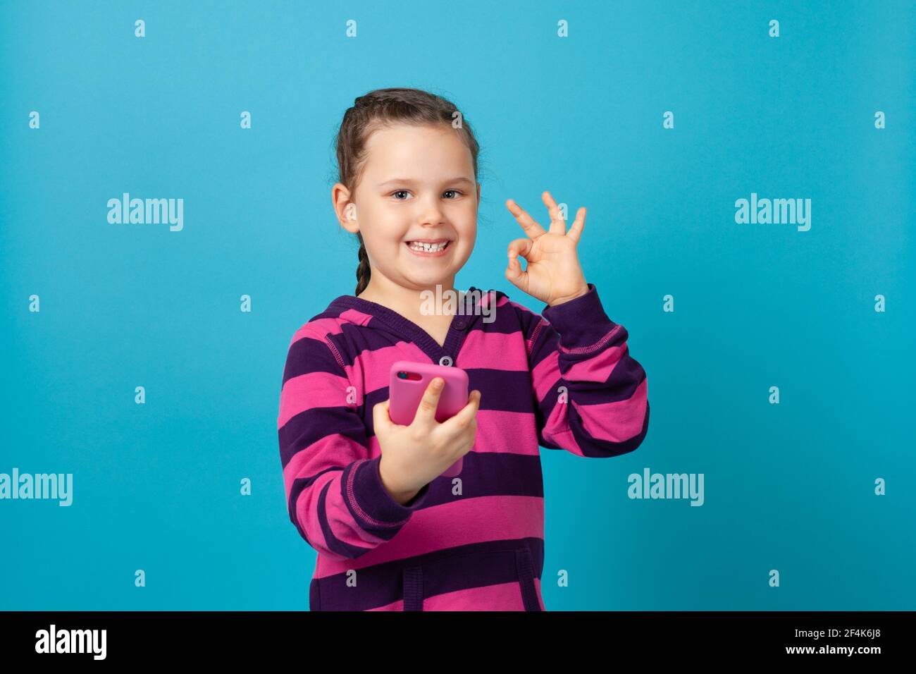 ritratto di una ragazza sorridente con i pigtail che tengono un telefono in un caso t rosa e mostrando un gesto ad anello isolato su sfondo blu Foto Stock