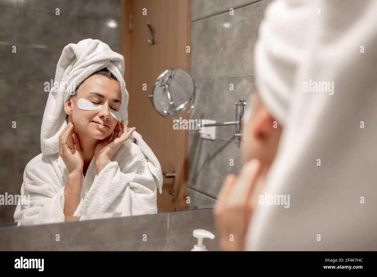 Una ragazza un accappatoio e con un asciugamano sulla sua testa bastoni macchie sotto i suoi occhi nel bagno di fronte allo specchio. Foto Stock