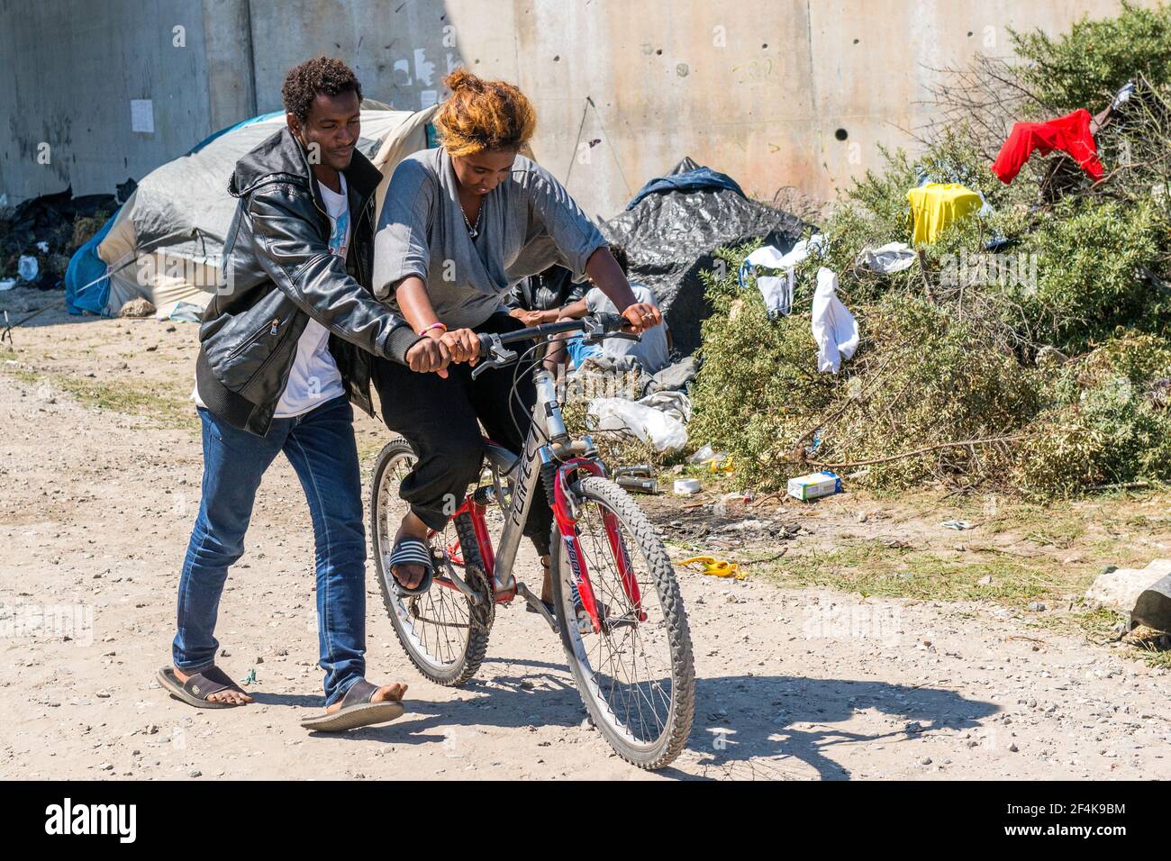 Calais, Francia. Un immigrato Eritreo illegale dal Continente Africano insegnando ad un collega migrante come guidare una bicicletta, mentre attendendo vicino ad una strada locale verso l'Oceano Atlantico / il Chanel verso il Regno Unito / Inghilterra. Lui e molti dei suoi colleghi rifugiati / migranti vivono in cattive condizioni all'interno della giungla, poiché le misure per impedire di viaggiare nel Regno Unito, rendono una transizione sempre più difficile. Foto Stock