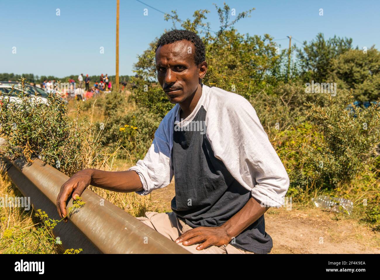 Calais, Francia. Un migrante illegale dal continente africano in attesa vicino ad una strada locale verso l'Oceano Atlantico / il Chanel verso il Regno Unito / Inghilterra. Lui e molti dei suoi colleghi rifugiati / migranti vivono in cattive condizioni all'interno della giungla, poiché le misure per impedire di viaggiare nel Regno Unito, rendono una transizione sempre più difficile. Foto Stock