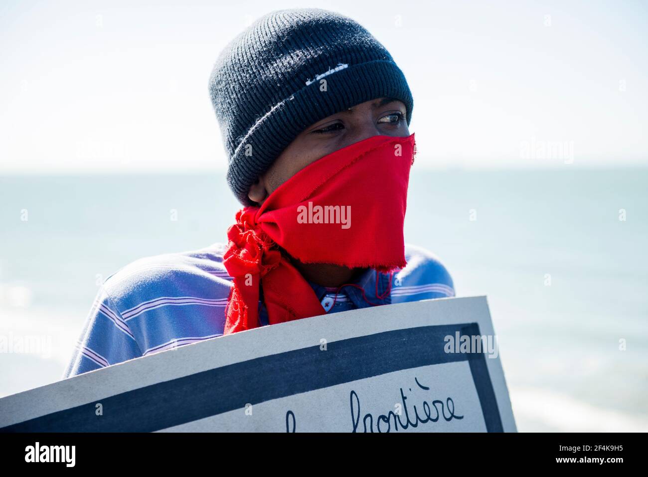 Calais, Francia. Un immigrato illegale dal continente africano che indossa un facemask e un segno di protesta mentre si affaccia sull'Oceano Atlantico / il Chanel verso il Regno Unito / Inghilterra. Lui e molti dei suoi colleghi rifugiati / migranti vivono in cattive condizioni all'interno della giungla, poiché le misure per impedire di viaggiare nel Regno Unito, rendono una transizione sempre più difficile. Foto Stock