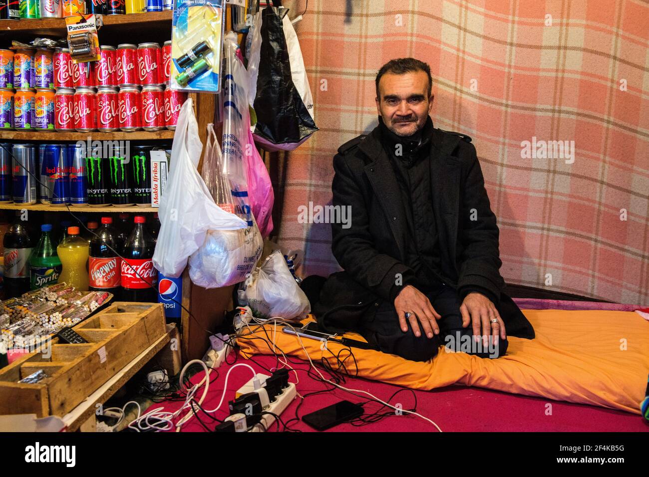 Calais, Francia. Mercante migrante illegale seduto nel suo negozio con merce, caricare smartphone a un punto di ricarica. Foto Stock