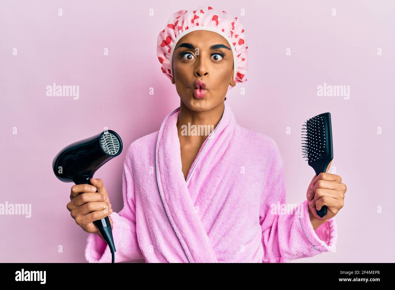 L'uomo ispanico che indossa la cuffia della doccia che tiene  l'asciugacapelli e il pettine che fa il viso di pesce con la bocca e gli  occhi squinting, pazzo e comico Foto stock -