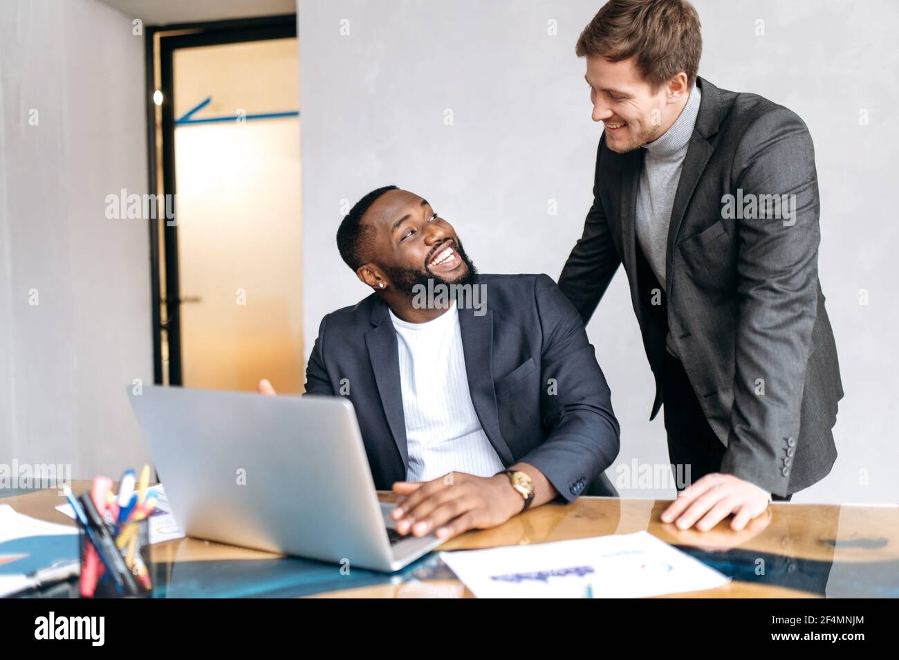 Le persone d'affari felici discutono le domande di lavoro in ufficio, usando il laptop, sviluppando insieme un nuovo progetto. Un impiegato africano americano maschio che chiede un consiglio da un collega caucasico uomo, sorridendo Foto Stock