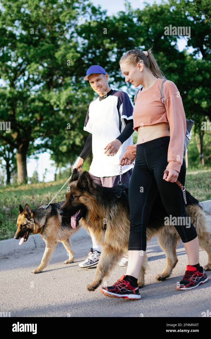 PET Love, addestramento del cane, migliori razze di cane per la famiglia. Giovani coppie sportive che camminano con due cani tedeschi Shepherd all'aperto nel parco estivo Foto Stock
