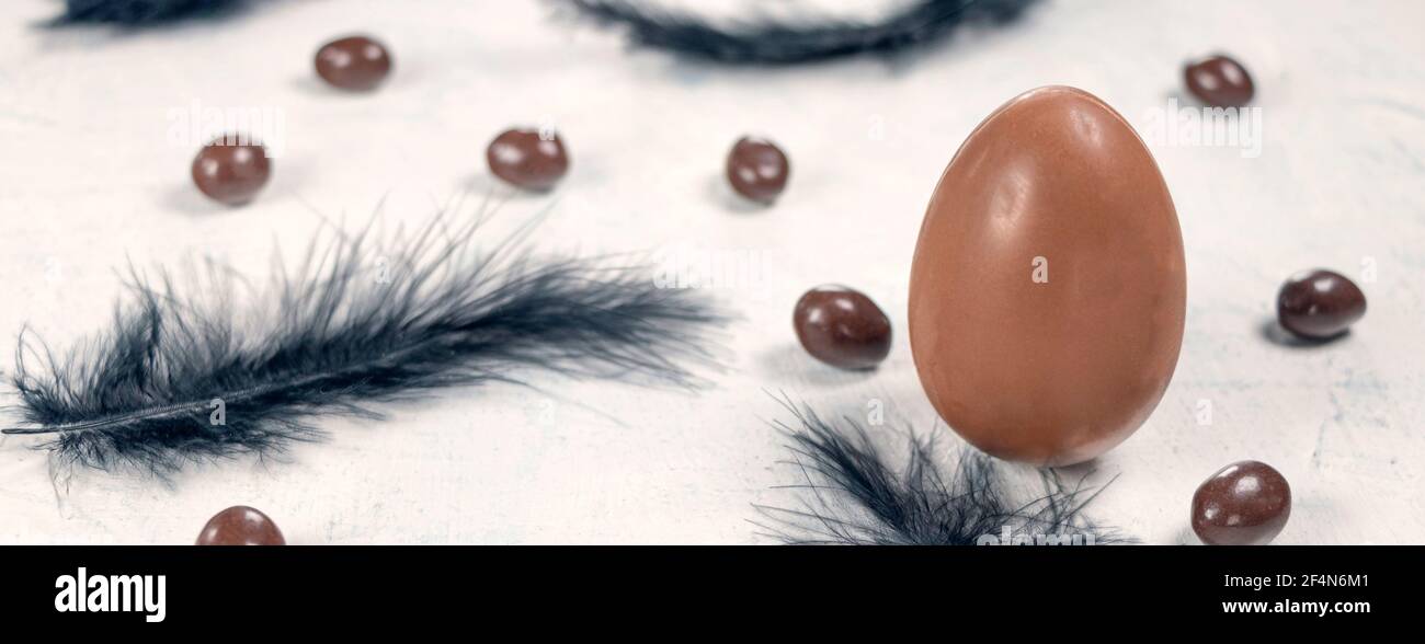 Uovo di cioccolato con piume nere e cioccolatini a forma di uovo su fondo bianco di cemento. Buon concetto di Pasqua. Foto Stock