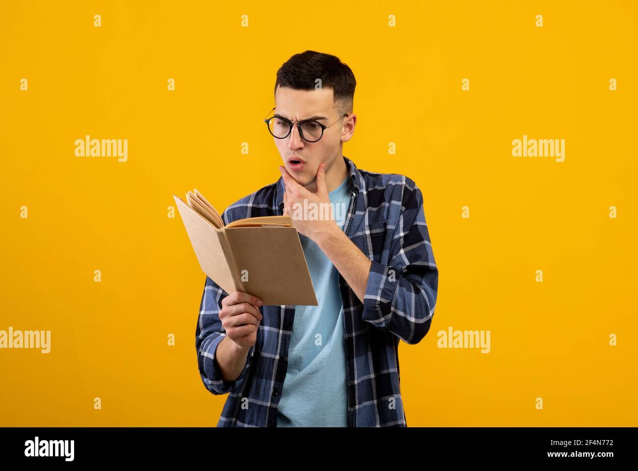Scioccato giovane studente in occhiali leggere libro di testo, avendo difficoltà a studiare per l'esame su sfondo studio arancione Foto Stock