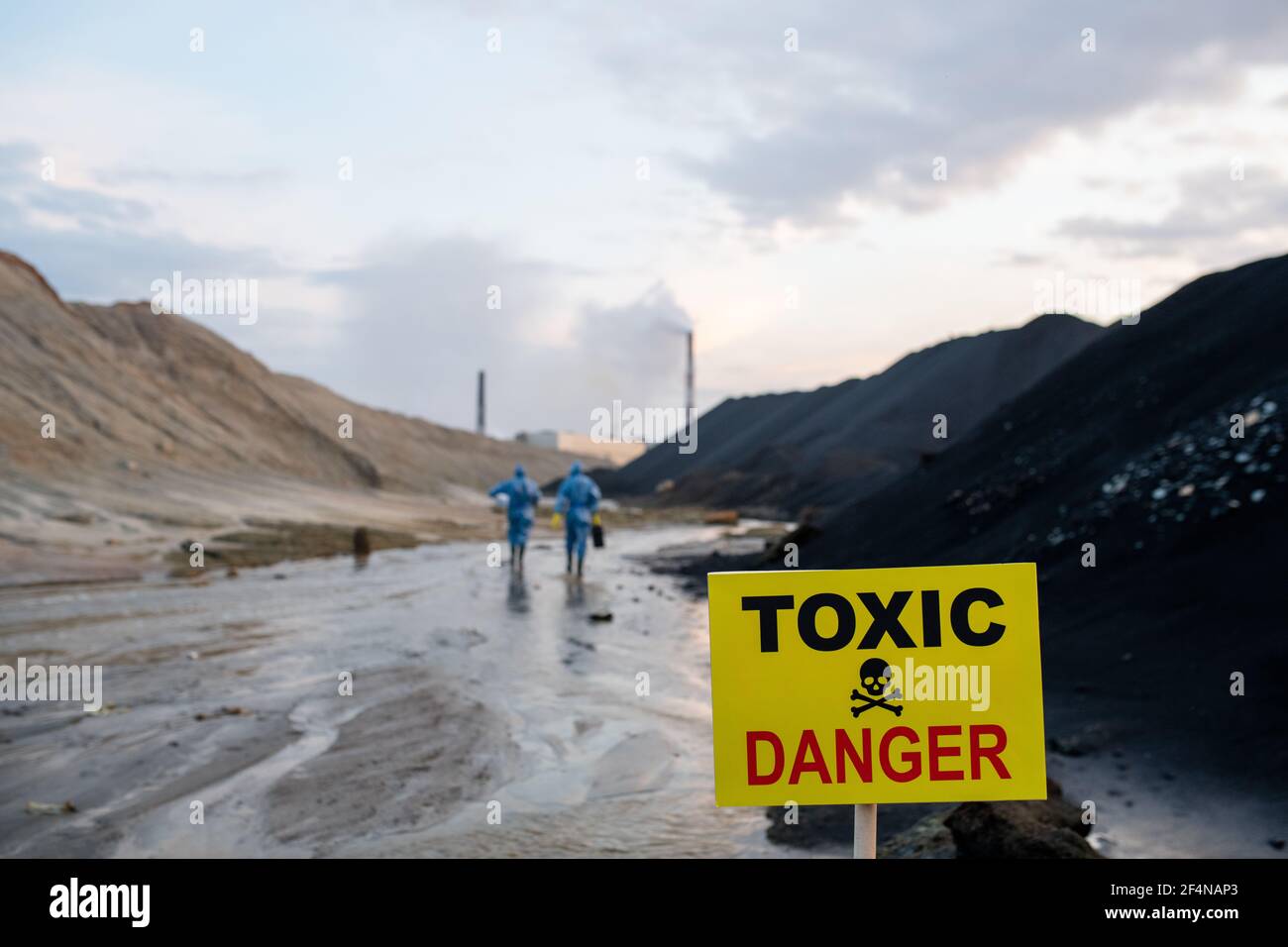 Cartoncino giallo con annuncio di area tossica e pericolosa su sfondo di due scienziati contemporanei in tute protettive blu Foto Stock