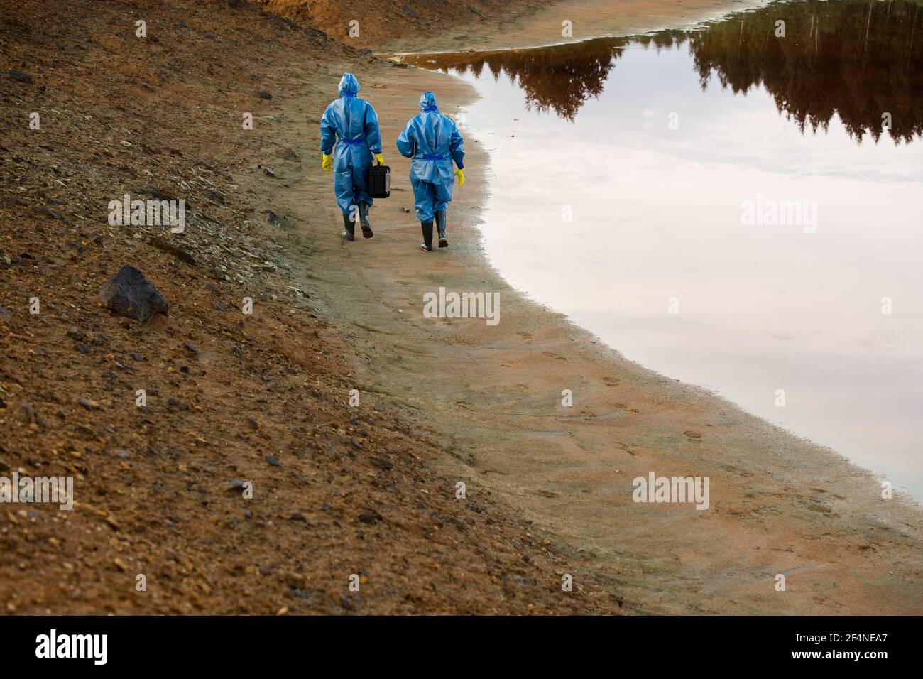 Vista posteriore di due ricercatori in tute che si muovono lungo il fiume con acqua sporca e suolo mentre effettuante l'indagine di il territorio Foto Stock