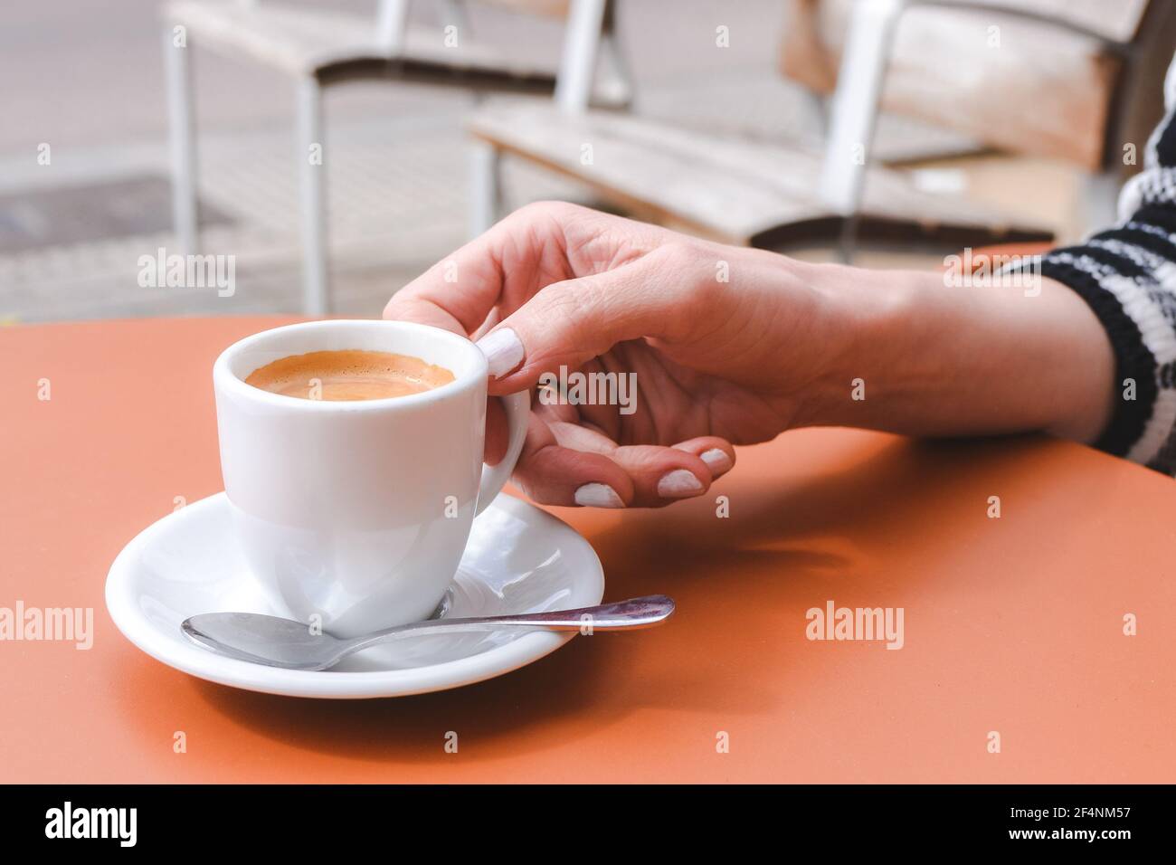 mano di una giovane donna caucasica che tiene una tazza bianca di caffè sulla parte superiore di un tavolo arancione a. una caffetteria con terrazza Foto Stock