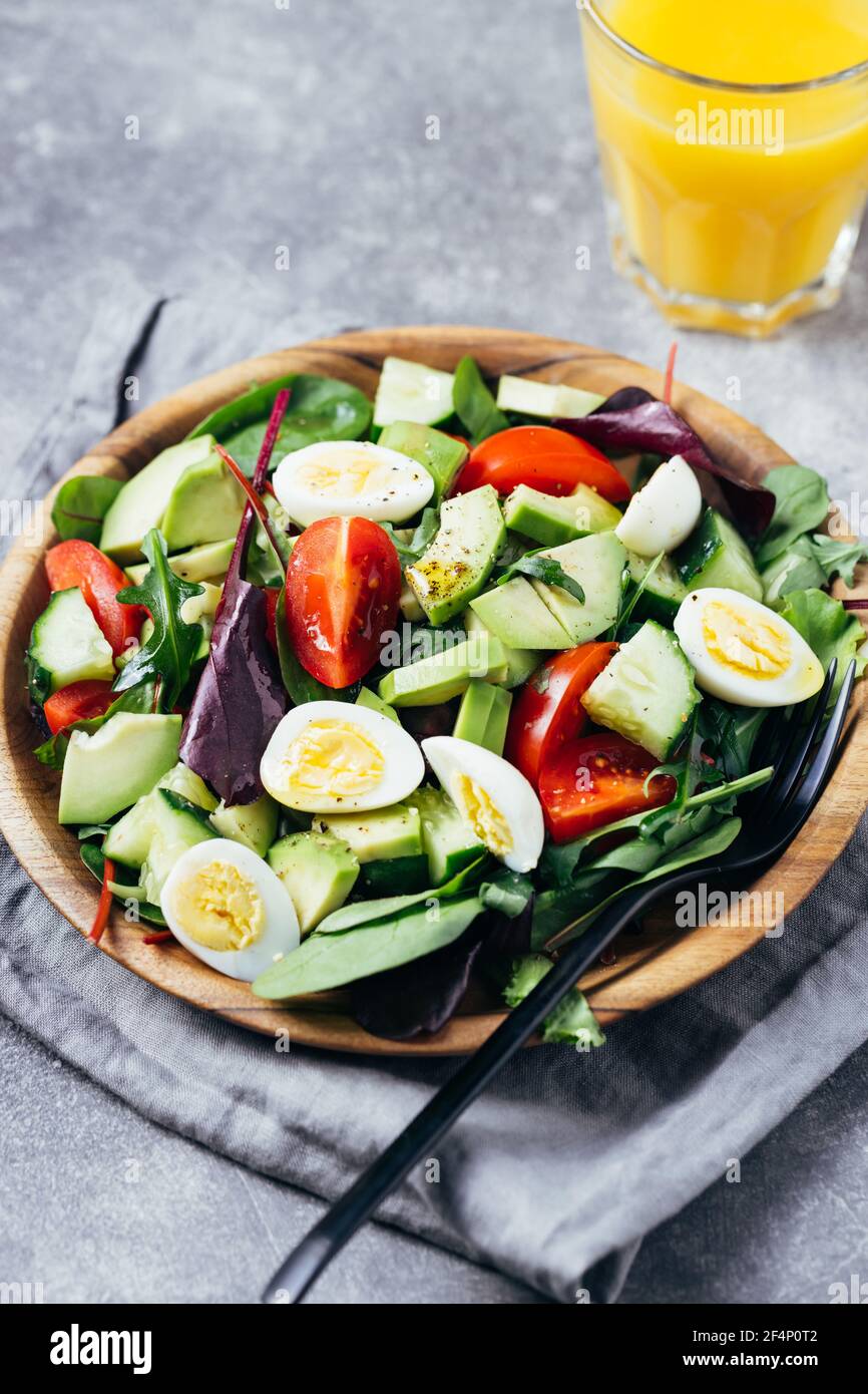 Insalata verde con pomodori, avocado, cetrioli e uova su piatto di legno e succo d'arancia sul tavolo. Foto Stock