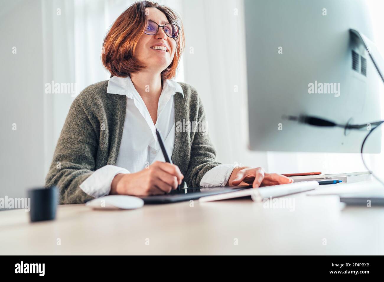 Donna sorridente sinceramente in occhiali utilizzando un tablet grafico con penna e computer moderno disegno grafica nel suo soggiorno. Ufficio domestico, distanza o. Foto Stock